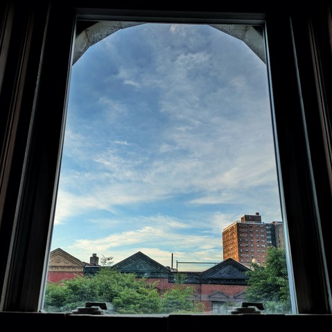 Looking through an arched window forty three minutes after sunrise the light blue sky is full of watery blue clouds. Pointed roofs of Harlem brownstones with red brickwork are across the street, and a taller apartment building can be seen in the distance. The green tops of two trees are starting to fill the frame on the bottom and right.
