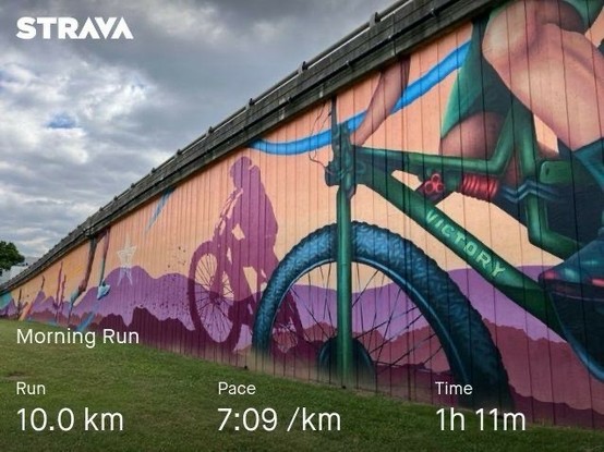 A mural of cyclists on a wall under a cloudy sky is shown. The Strava app overlay displays details of a 10.0 km morning run with a pace of 7:09/km and a duration of 1 hour and 11 minutes