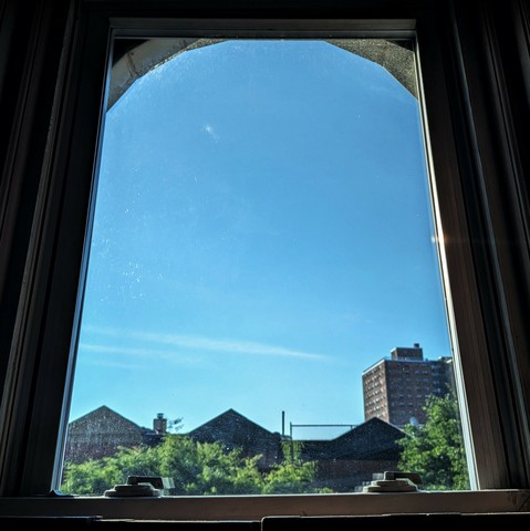 Looking through an arched window two hours after sunrise the blue sky is crossed with two subtle lines of cloud. Pointed roofs of Harlem brownstones are silhouetted across the street, and a taller apartment building can be seen in the distance. The green tops of two trees are starting to fill the frame on the bottom and right.
