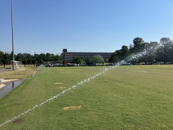 A grassy sports field with sprinklers activated, spraying water.