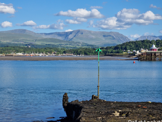 Snowdonia mountains in the distance over the Menai Straits. Bangor Pier sits across the water. It is sunny.