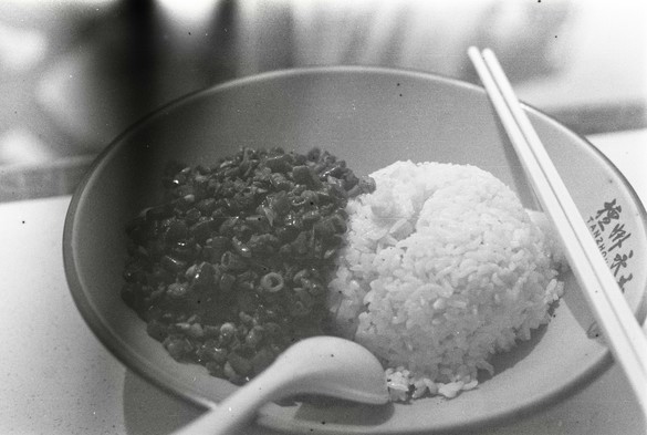 A black and white image of a bowl of steamed rice with some fried beans and minced meat. A pair of chopsticks lie on top of the bowl to the right.