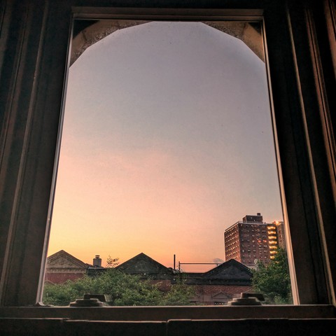 Looking through an arched window twenty minutes before sunrise the sky is suffused with orange and pink mist. Pointed roofs of Harlem brownstones with red brickwork are silhouetted across the street, and a taller apartment building can be seen in the distance. The green tops of two trees are starting to fill the frame on the bottom and right.