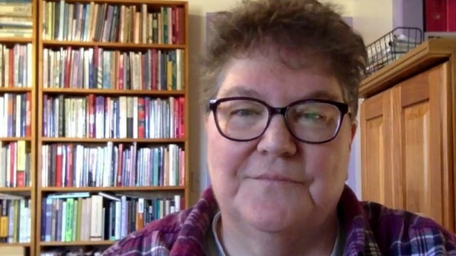 A woman with short cut hair and dark rimmed glasses standing in front of bookshelves filled with books.