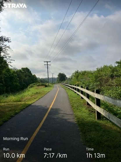 A paved trail surrounded by greenery, power lines, and a fence on one side. Overlay text shows a 10 km morning run with a pace of 7:17 per km and a time of 1 hour and 13 minutes.