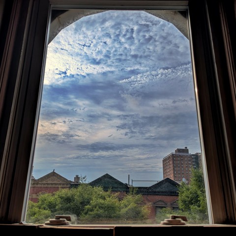 Looking through an arched window two hours after sunrise the blue sky is full of fine, dense, mottled white clouds. Pointed roofs of Harlem brownstones with red brickwork are across the street, and a taller apartment building can be seen in the distance. The green tops of two trees are filling the frame on the bottom and right.