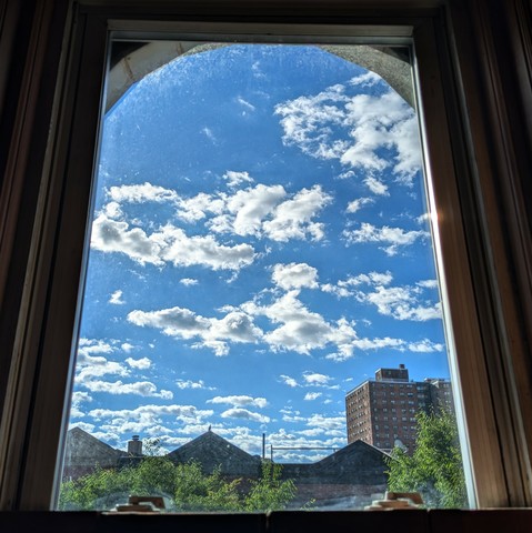 Looking through an arched window two and a half hours after sunrise the bright blue sky is full of scattered horizontal white puffy clouds. Pointed roofs of Harlem brownstones are silhouetted across the street, and a taller apartment building can be seen in the distance. The green tops of two trees are filling the frame on the bottom and right.