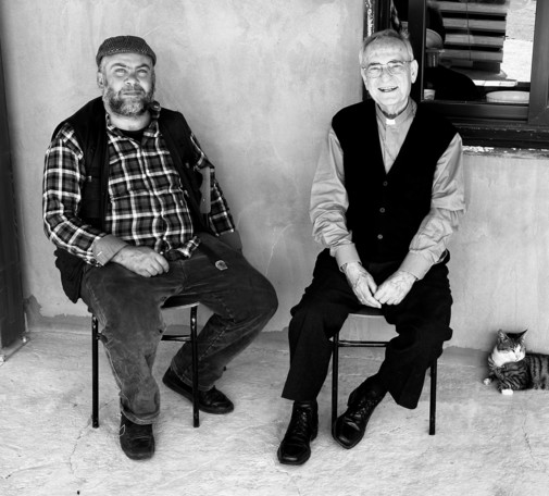 A black & white photograph of two members of the clergy. They sit on chairs in the open shade of a farm house veranda. The man on the left is also a farmer and is dressed in clothes more suited to farming. A cat reclines on the floor, partly out of the frame, next to the man on the right.