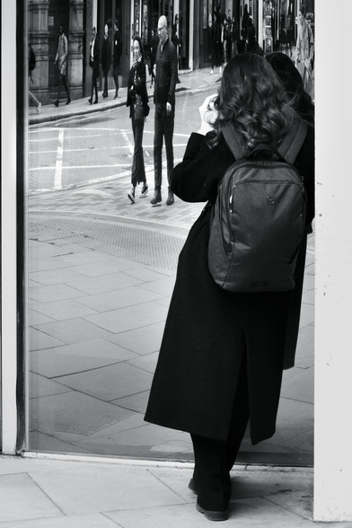 A black and white photo of a lady photographer, all in black and with a matching backpack, leaning in a doorway and taking photos in the mirror on the door, reflected in which are two people, squashed thin by the distorted glass.