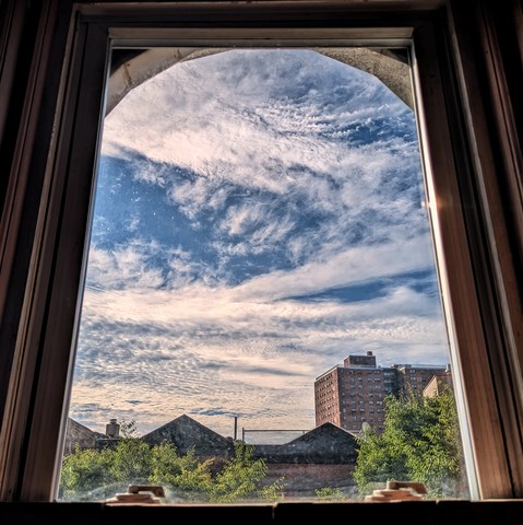 Looking through an arched window two hours after sunrise the blue sky is full of swirling high-altitude white clouds. Pointed roofs of Harlem brownstones are across the street, and a taller apartment building can be seen in the distance. The green tops of two trees are filling the frame on the bottom and right.