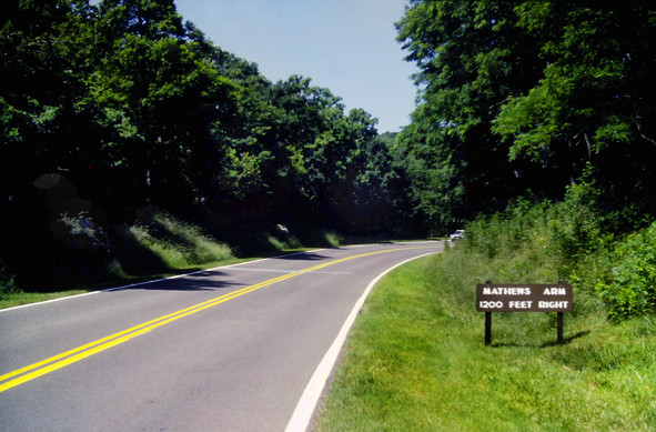 Crossing the two-lane park road, Skyline Drive. Its narrow grassy shoulders are surrounded by forest. A car is heading towards us, just coming around a bend up ahead. To our right is a rustic wooden sign reading in all caps, 