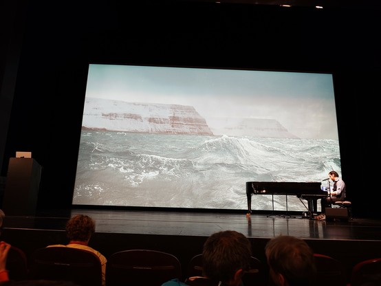 The stage has a huge projection of a snowy promontory in a very stormy sea. To the right, a pianist is playing at a concert piano.