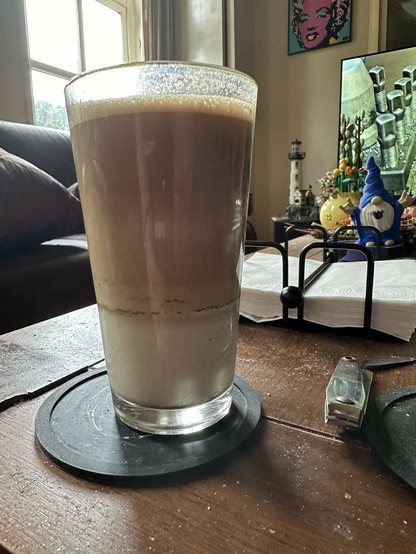 The image shows a tall glass filled with a layered beverage, likely a latte or similar drink. The bottom layer is white, the middle layer is a light brown, and the top layer is a darker brown, indicating different densities of milk and coffee. The glass is placed on a black coaster on a wooden table. In the background, there is a couch near a window, a colorful piece of artwork on the wall, and a TV screen showing an image of bolts or screws. There is also a small blue gnome figurine and a lighthouse decoration on the table, along with a metal nail clipper.