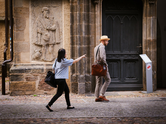 Ein Mann in elegeantem Someroutfit geht an einer Kirchentür vorbei. Hinter ihm ist eine Frau, die auf ihn zu zeigen scheint. 