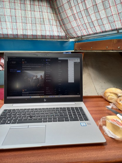 A laptop on a small table inside a boat. Checkered curtains are closed infront of the windows. To the right of the laption there's some cheese and a bag of buns.