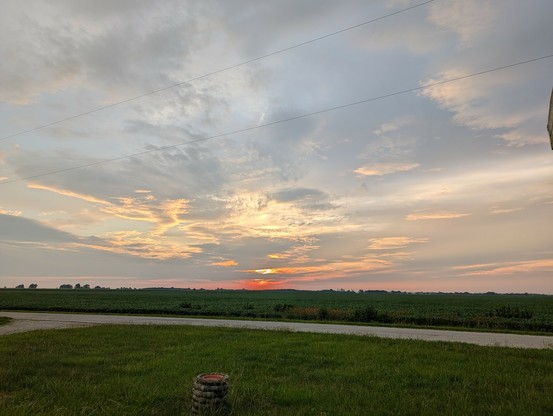 Sunrise over the corn field. Cloudy today, rain and hurricane leftovers moving in. The sun is just over the horizon, and there is a big orange spot where it's trying to break through the clouds. There are some spots of silver and peach in the clouds above.