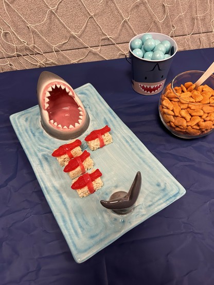 Ceramic shark cutting board with sushi made from Rice Krispie treats and Swedish fish. A bowl of goldfish crackers is on the right. 