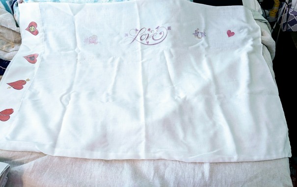 A vintage white pillow case. It has hearts of red with different designs on the edge. More faded are a Cupid, 