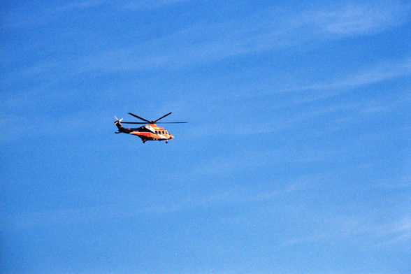 An orange and black helicopter hangs in a blue and whisp cloud sky. The blades appear frozen in place. 