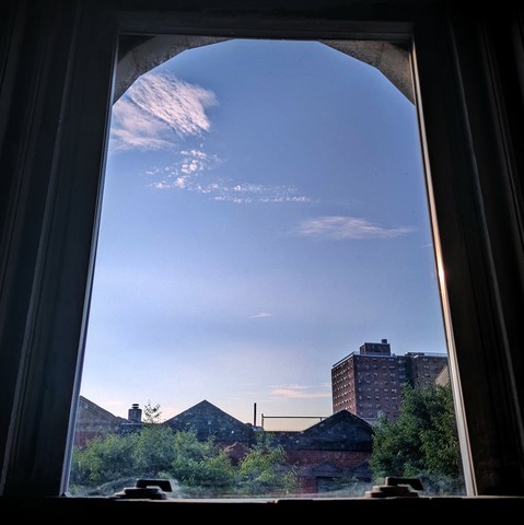Looking through an arched window two hours after sunrise the pale blue sky is marked with a few spiny hieroglyphic clouds. Pointed roofs of Harlem brownstones with red brickwork are silhouetted across the street, and a taller apartment building can be seen in the distance. The green tops of two trees are filling the frame on the bottom and right.