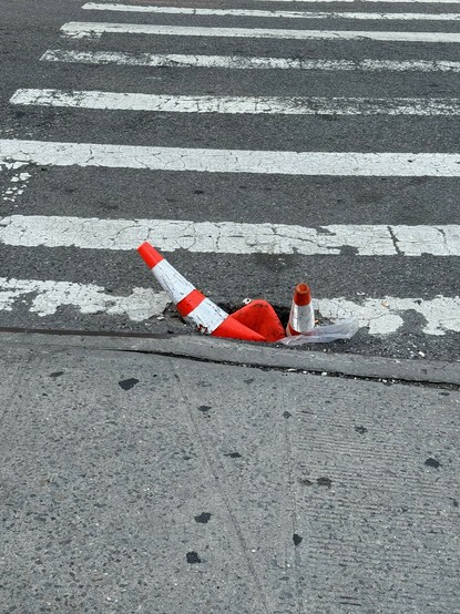A pothole with two orange cones inside of it, sticking out of the street at a crosswalk,  near a curb