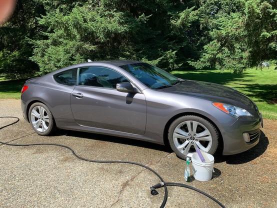 A silver Genesis Coupe shown from the side, having just been washed. A bucket and hose are near the front wheel. It's parked on a driveway with fir trees and grass in the background. It's a sunny day for a car wash.