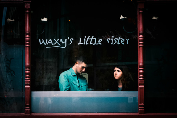 A cinematic colour photo of two young people, a man and a woman, sitting in the window of a bar called Waxy's Little Sister.