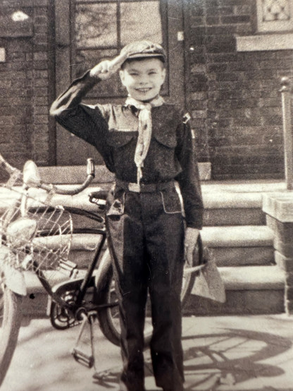 Photo of an 8 year old white boy (c’est moi) standing in front of the stoop of his house. Behind him is the bicycle he just got for his birthday. He’s wearing his Cub Scout uniform, proudly giving the official two-fingered salute. 1958.