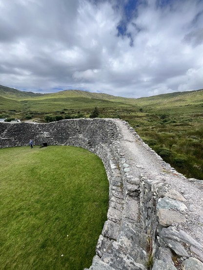 A fortress wall circa 5th-11thC. Dry stone walled grey stones in a circle with steps internally to reach the top. Green mountains and blue skies behind