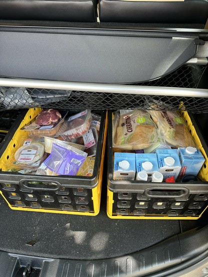 The image shows the back of a car with the trunk open. Inside the trunk, there are two yellow and black plastic crates filled with groceries. 

The crate on the left contains various food items, including packaged meat, cheese, and other groceries. The crate on the right contains several cartons of milk and packages of bread. The crates are placed side by side, and there is a netted barrier above them, likely to prevent items from shifting during transport. The back seats of the car are visible above the netted barrier.