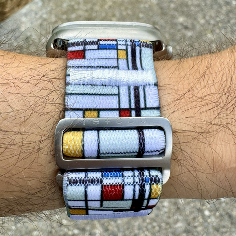 A closeup of a textile-textured Apple Watch band with mat silver metal buckle. The band is predominantly white with rectangles of colour