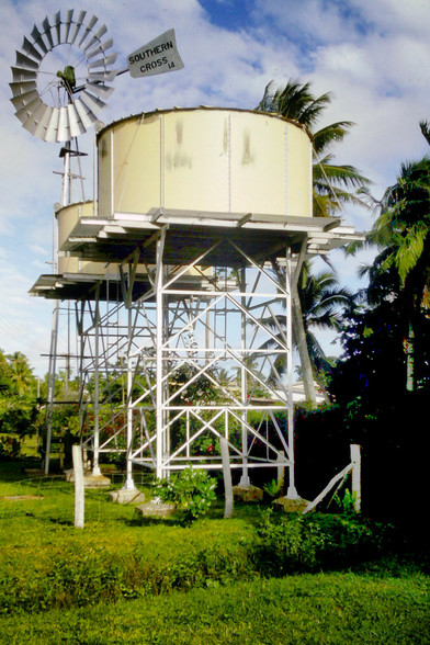 Beneath a partly cloudy sky, two pale yellow metal water tanks and a windmill rise from a lush grassy area surrounded by palm trees and other tropical vegetation. The tanks are raised off the ground a few dozen feet by white scaffolding, the windmill stands slightly higher. It is labeled 