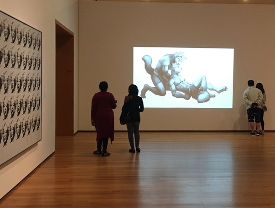 Museum gallery with people viewing art. Anthropomorphic-themed image projected on the right wall, and repeated black-and-white faces of Marilyn Monroe framed on the left.