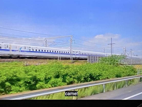 Still photograph from a Japanese travel video:

A Shinkansen (bullet train) passing at speed, seen through the window of a vehicle on a road that runs parallel to the railway line.

Closed caption at the bottom of the screen:

