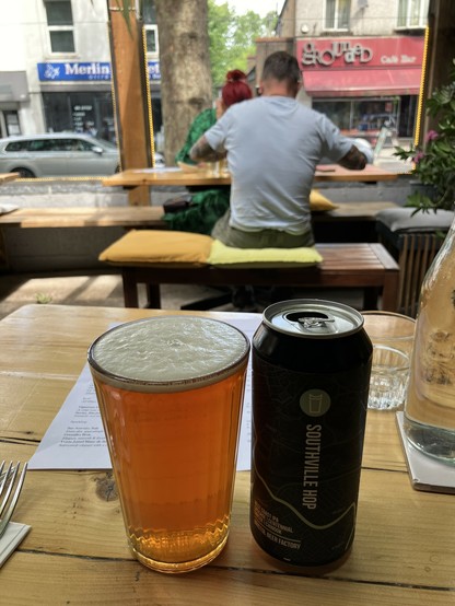 A pint of coppery beer, poured from a can, sitting on a wooden table on a restaurant terrace, with a view of an English high street in the background. The dark, stylish can says 