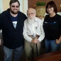 A man (Bob) and a woman (Eve) flank a little old bearded man with a cane (James Randi)