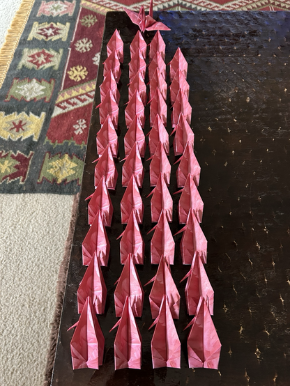 40 bright pink origami cranes on a dark wooden table. 39 are folded, and 1 is opened.