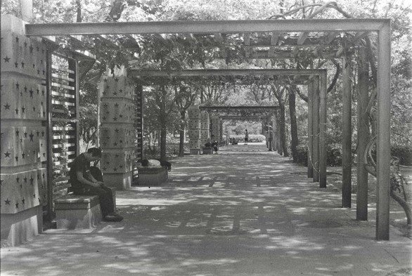 A black and white image of people napping and sitting in a passage formed by steel pillars attached to a side of the wall in the shades in a park.
