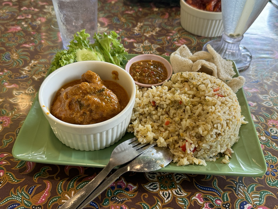 A plate of chili padi fried rice and ayam rendang. The dish includes a serving of spicy fried rice with red chilies, a bowl of ayam rendang (chicken in a rich, spiced coconut milk stew), fresh lettuce and cucumber, a small bowl of sambal, and prawn crackers.