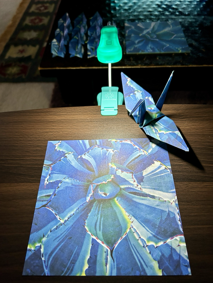 Photo of an opened origami crane on a wooden desk, along big with an unfolded sheet and some folded yet unopened ones in the background. The print of the paper is a bluish multicolor cactus.
