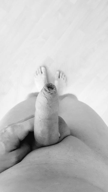 Pov and greyscale photo of my naked underside looking down towards my bare feet, where I hold my soft and foreskin covered penis. No pubes