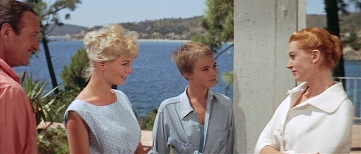 The image depicts four individuals standing outdoors near a body of water, likely a sea or lake, with a scenic background of trees and distant hills. The person on the far left, dressed in a reddish shirt, stands next to a young blonde woman in a light blue, sleeveless polka-dot dress. Next to her is another young woman with short blonde hair, wearing a light blue button-up shirt. On the far right, a woman with red hair is dressed in a white outfit with a large collar. The group appears to be engaged in a conversation. Behind them, there are green plants and a clear view of the water and the distant landscape.