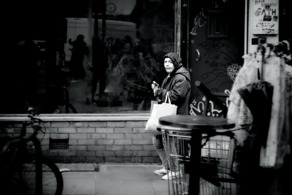 A high-contrast black and white photo taken with a vintage lens of an old lady walking through London's Brick Lane Market with something she's bought, clutching it in both hands. Just as I took the photo she glanced over at me.
