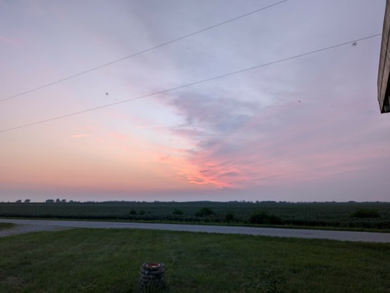 Sunrise over the corn field. A departing thunderstorm is covering the right half of the sky,the left is light blue and clearing. There's a little bit of pastel pink at the bottom of the storm.
