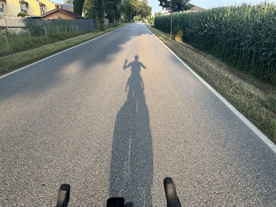 Ein Schatten von mir auf dem Fahrrad auf einer Asphaltstraße, der mit einer Hand winkt.