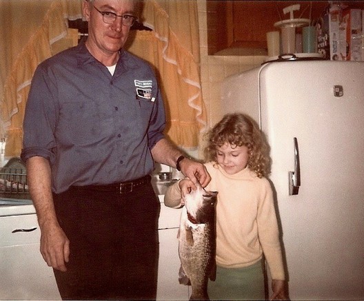 Me and my Dad. A Caucasian man on the left wearing glasses his blue shop uniform from the Ford/Mercury dealership. On the right, me, a caucasian young girl wearing a yellow sweater and green pants, blonde curly hair. They are together holding a large fish, which was about to be dinner that was just caught on a trip to a local fishing hole. The photo takes place in my childhood kitchen with white metal sink area, white refrigerator, frilly apricot curtains, plywood cupboards, tuperware cup set, cereal boxes. 