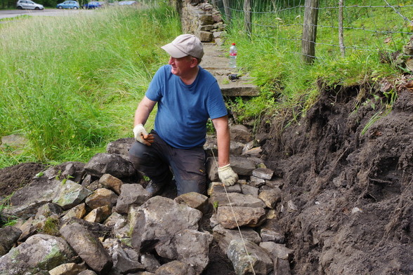 A man kneels in between a pile of stones and the wall he is rebuilding. He wears a blue t shirt, grey cap, orange gloves and black work trousers. He is smiling and listening to someone out of shot to his right. An already part repaired length of wall extends behind him. 