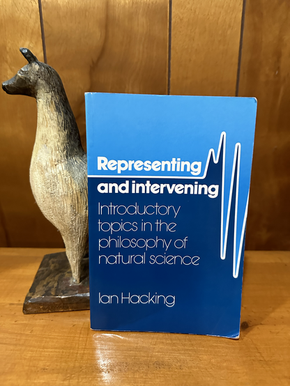 A book resting against a llama carved in wood, on top of a wood surface, backed by a fake-wood (very 1970s) wall paneling. 

The title is /Representing and Intervening: Introductory Topics in the Philosophy of Natural Science/, by Ian Hacking.

The cover is too boring to describe. There’s light blue on the top, darker blue on the bottom.