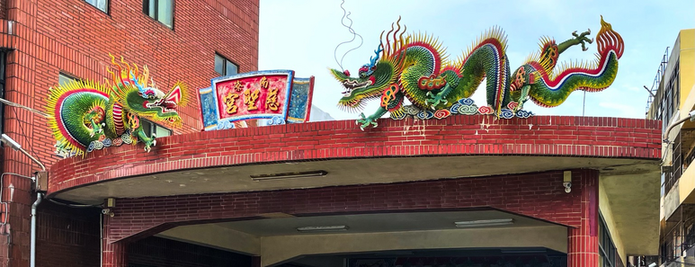 Two mostly green Chinese dragons face each other across the temple sign, atop a red tile flat roof building. A red brick apartment building in the background. 