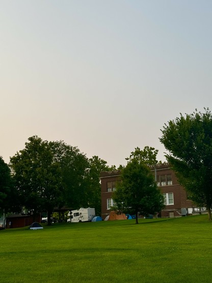 Grassy field with trees, tents, an RV, and a brick building in the background under a clear sky.